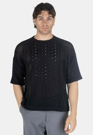 Uomo che indossa un maglione nero a maniche corte in maglia con motivo traforato e pantaloni grigi, in piedi con una mano in tasca su sfondo bianco.