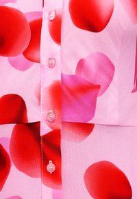 Κοντινό πλάνο σε ροζ ύφασμα με μεγάλες κόκκινες και μοβ αφηρημένες κηλίδες και διαφανείς κουμπιά κατά μήκος μιας ραμμένης ραφής.