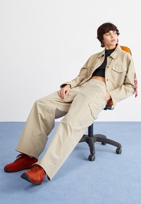 Beige cargo kalhoty s elastickým pasem a postranními kapsami, kombinované s černým crop topem a červenými sportovními botami. Sedí na otočné židli.