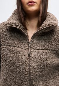 Γυναίκα που φοράει ταμπά τζάκετ από φλις με υφή, με ψηλό γιακά και φερμουάρ μπροστά που είναι μερικώς κλειστό.