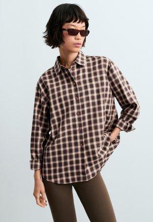 Woman wearing dark sunglasses and a brown and beige plaid button-up shirt with sleeves rolled up, posing with one hand in pocket.