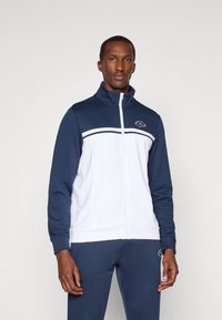 Mann, der eine navyblaue und weiße Sportjacke mit Reißverschluss und passende Hose mit Logo auf der Brust und dem Oberschenkel vor einem schlichten Hintergrund trägt.