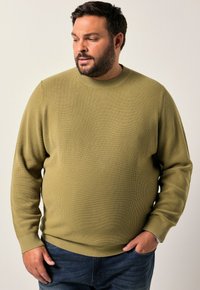 Maglione a coste verde oliva con collo rotondo e maniche lunghe, abbinato a jeans scuri. Il tessuto ha un aspetto strutturato.