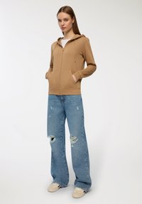 Felpa beige con zip e tasca anteriore, abbinata a jeans strappati azzurri chiari e sneaker beige, su uno sfondo chiaro e uniforme.