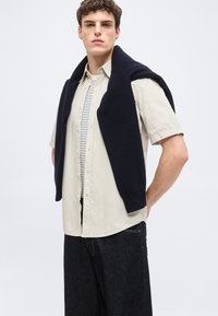 Άνδρας που φοράει μπεζ πουκάμισο με κοντά μανίκια πάνω από ριγωτό μπλουζάκι, σκούρο τζιν και ναυτικά πουλόβερ περασμένο στους ώμους, στέκεται μπροστά σε απλό φόντο.