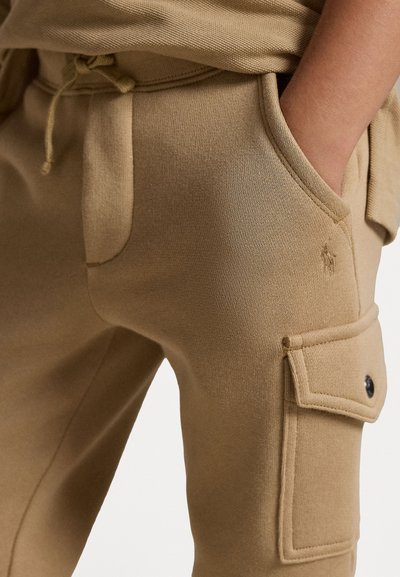 Calças de algodão bege com cintura elástica, cordão, bolsos laterais e bolso cargo com fechamento por botão; textura suave, design ajustado.