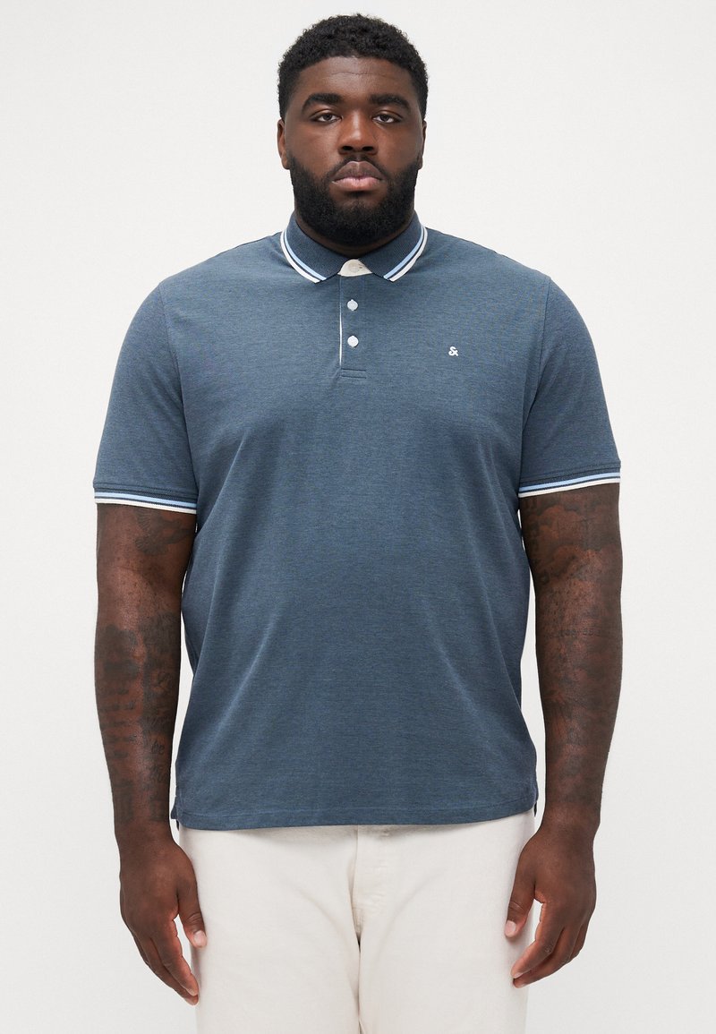 Man met tatoeages draagt een blauwe polo met korte mouwen en een witte gestreepte kraag en mouwen, gecombineerd met lichtbeige broek, staand tegen een witte achtergrond.