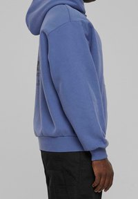 Poloje i mjukt lavendelfärgat tyg, med en känguruficka och ribbade ärmslut. Designen inkluderar tryckt text på baksidan.