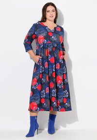 Robe en coton avec un col en V, des manches 3/4 et une silhouette évasée. Présente un fond bleu marine avec des motifs floraux rouges et bleus.