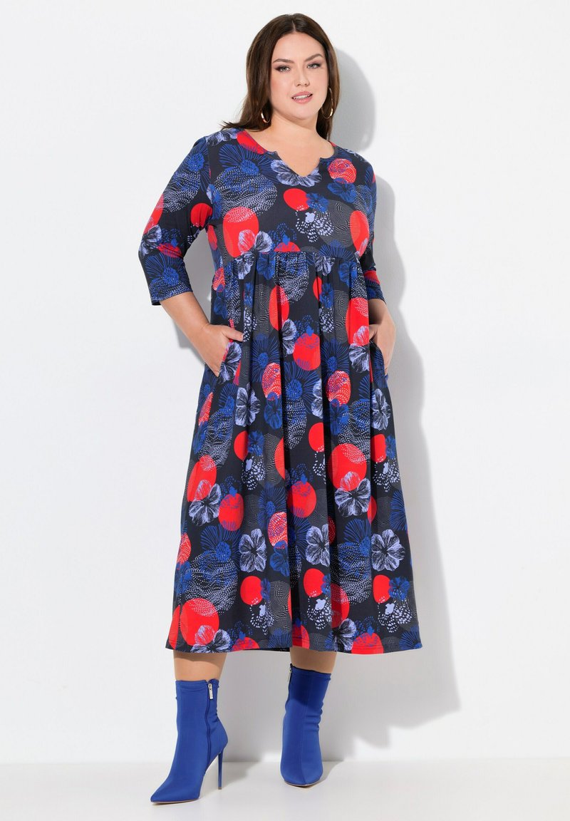 Robe en coton avec un col en V, des manches 3/4 et une silhouette évasée. Présente un fond bleu marine avec des motifs floraux rouges et bleus.