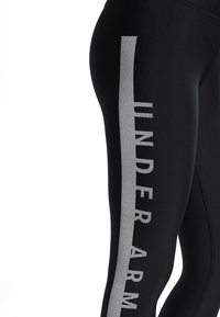 I leggins neri presentano una striscia verticale lucida e ventilata con il testo "UNDER ARMOUR" in grigio lungo il lato per il branding e la visibilità.