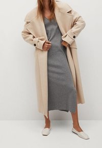 Femme portant un long manteau beige sur une robe midi grise et des ballerines blanches, se tenant contre un mur blanc uni.