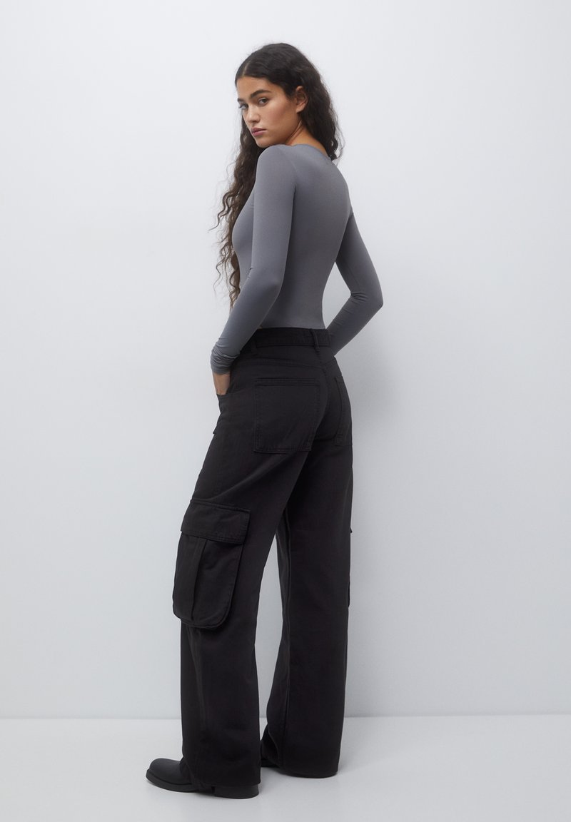 Maglia grigia a maniche lunghe e aderente abbinata a pantaloni cargo neri con tasche laterali, gamba larga e finitura opaca. Tessuto dalla texture liscia.
