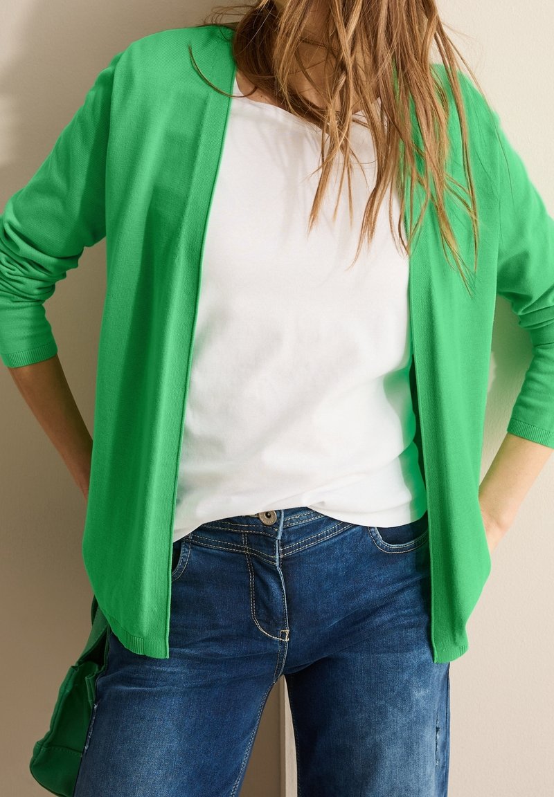 Personne portant un cardigan vert décontracté ouvert sur une chemise blanche et un jean bleu, debout avec les mains sur les hanches devant un fond uni.