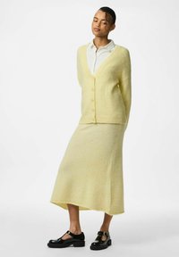 Geel gebreid vest met een V-hals en knoopsluiting, gecombineerd met een bijpassende lange rok. Zwarte gepatenteerde leren schoenen maken de outfit compleet.
