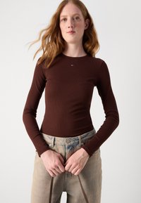 Hnědá dlouhá rukávová ribbed košile s malým vyšitým logem. Model je v světle modrých vysokých džínách a drží popruhy.