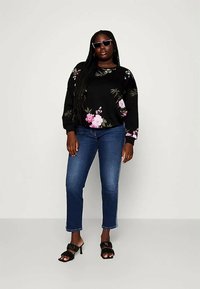 Mulher vestindo um sweatshirt preto com flores, jeans azuis, sandálias de salto pretas e óculos de sol escuros, de pé contra um fundo simples.