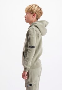 Olijfgroene hoodie van zacht materiaal, met een voorkant zak, een capuchon met trekkoord en een merkembleem op de mouw. Relaxed fit.