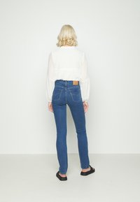 Blusa branca transparente com mangas bufantes, combinada com jeans azuis de cintura alta com bolsos traseiros e um patch de couro castanho. Sapatos pretos tipo slip-on.