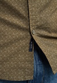 Camisa marrón con patrón de diamantes, cierre de botones y una etiqueta lateral que dice "PETROL IND." Combinada con jeans de mezclilla azules.