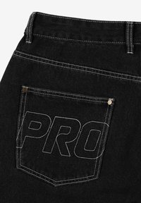 Czarne dżinsy mają wyraźnie wyszywaną tylną kieszeń z napisem "PRO", podkreśloną białą nitką i metalowymi nitami.