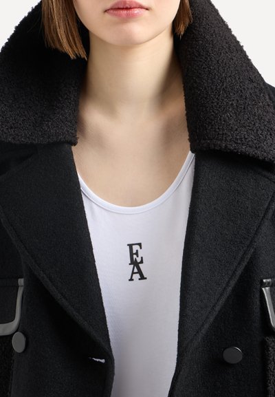 Cappotto di lana nera con colletto texturizzato, abbinato a una camicia bianca con il logo 'EA' nero. I bottoni sono rotondi e scuri.