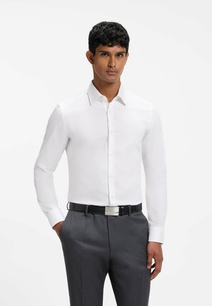 Mann trägt ein weißes, langärmliges Hemd mit Knöpfen, graue Hose und schwarzen Gürtel, steht mit einer Hand in der Tasche vor weißem Hintergrund.