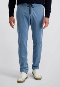 Man met lichtblauwe corduroy broek met zwarte trekkoord en witte sneakers, staand tegen een effen lichte achtergrond.