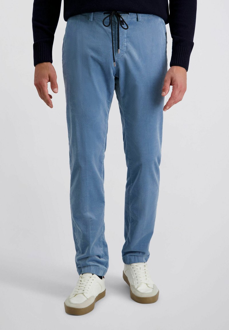 Man met lichtblauwe corduroy broek met zwarte trekkoord en witte sneakers, staand tegen een effen lichte achtergrond.