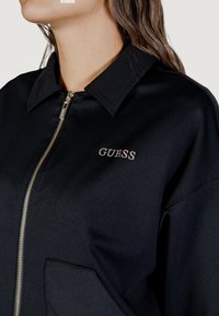 Giacca nera con zip e colletto, caratterizzata da un logo GUESS argento e una grande tasca applicata sul davanti. Tessuto morbido e strutturato.