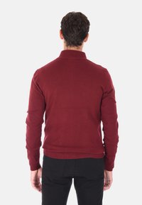 Maglione rosso lavorato a maglia con colletto e polsini a costine, caratterizzato da un design aderente. Indossato con pantaloni neri, mostrando una texture liscia.