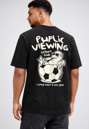 Mann trägt ein schwarzes T-Shirt mit einem Cartoon-Vogel, der in einem Fußball entspannt liegt, und Text "Public Viewing Leisure Club Living easy is the goal".