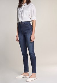 Jeans de tiro alto de color azul oscuro con un ajuste slim, combinados con una camisa blanca de botones. El conjunto incluye zapatos slip-on blancos.
