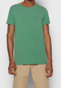T-shirt en coton vert avec un col rond, des manches courtes et un petit logo sur la poitrine gauche. Texture lisse, coupe décontractée.