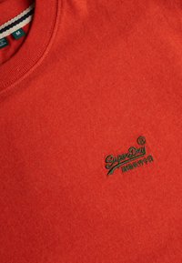 Zbliżenie czerwonej bluzy z okrągłym dekoltem z zielonym wyszywanym logo "SuperDry" oraz małymi azjatyckimi znakami na piersi.