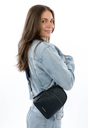 Noorpruunide pikkade juustega noor naine helesinises teksajakis ja teksades, naeratades, kandes õlal väikest musta ristiõlakotti.