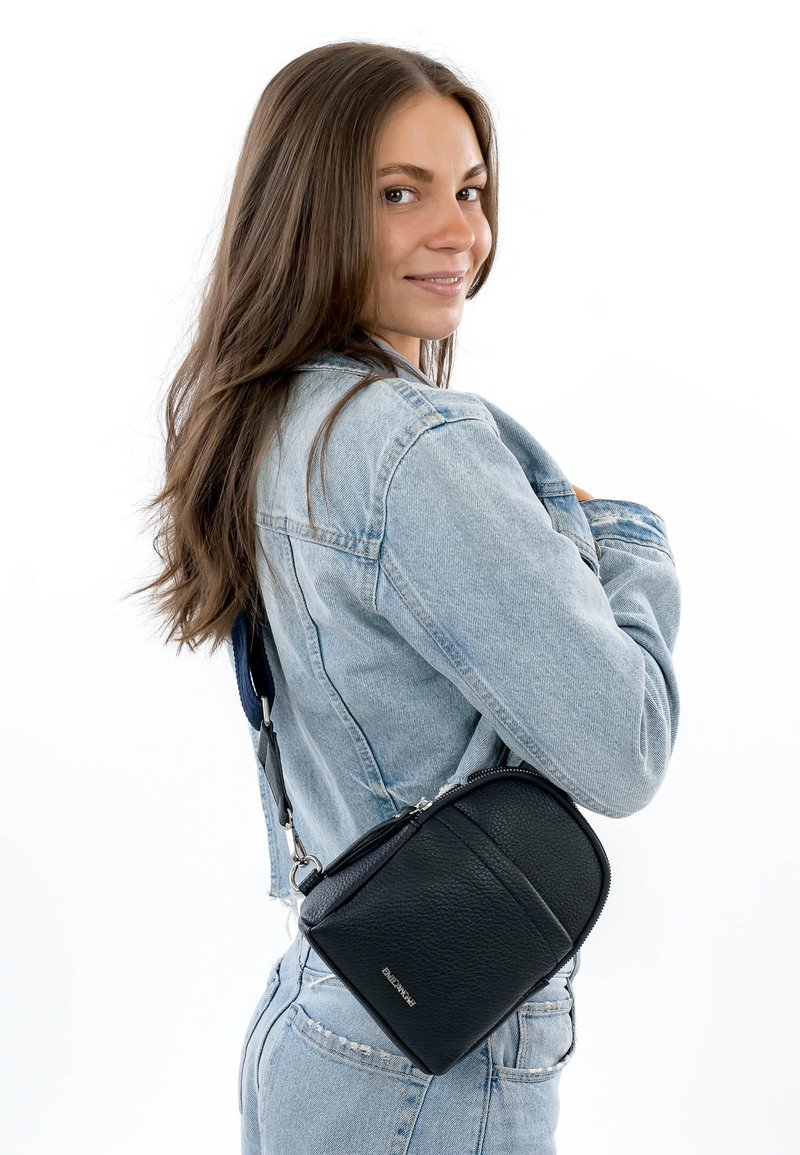 Junge Frau mit langen braunen Haaren in heller Jeansjacke und Jeans, lächelnd, mit einer kleinen schwarzen Umhängetasche über der Schulter.