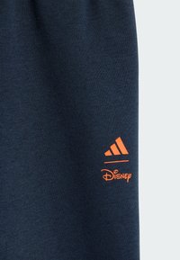 Tums zila sporta bikses ar gludu tekstūru, kurām ir oranžs Adidas logo un Disney zīmols tuvu apakšmalai.
