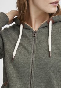Zelená mikina na zip s krémovými stahovacími šňůrkami, hnědými akcenty a texturovanou podšívkou. Obsahuje přední zip a žebrované manžety.