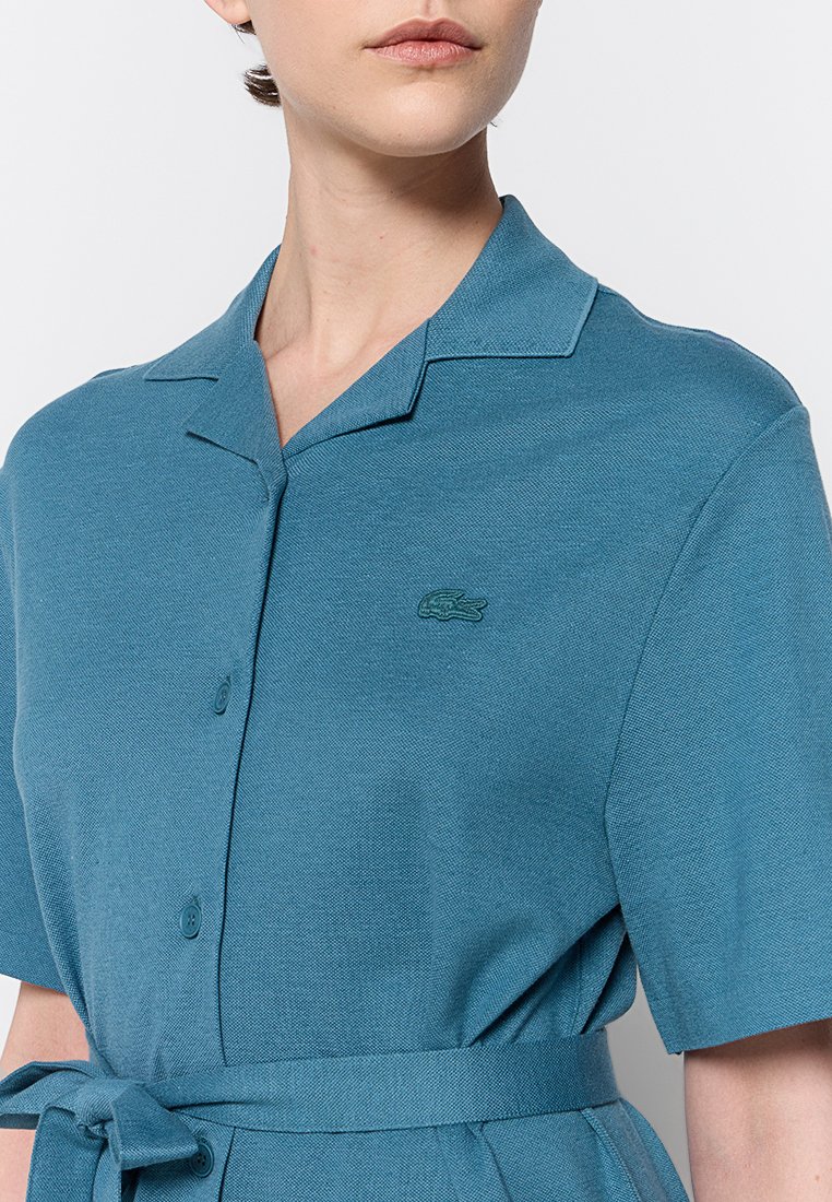 Kobieta ubrana w turkusową sukienkę z krótkim rękawem, z kołnierzykiem, zapinaną na guziki, z wiązaną w talii paskiem, z małym haftowanym logo krokodyla na piersi.