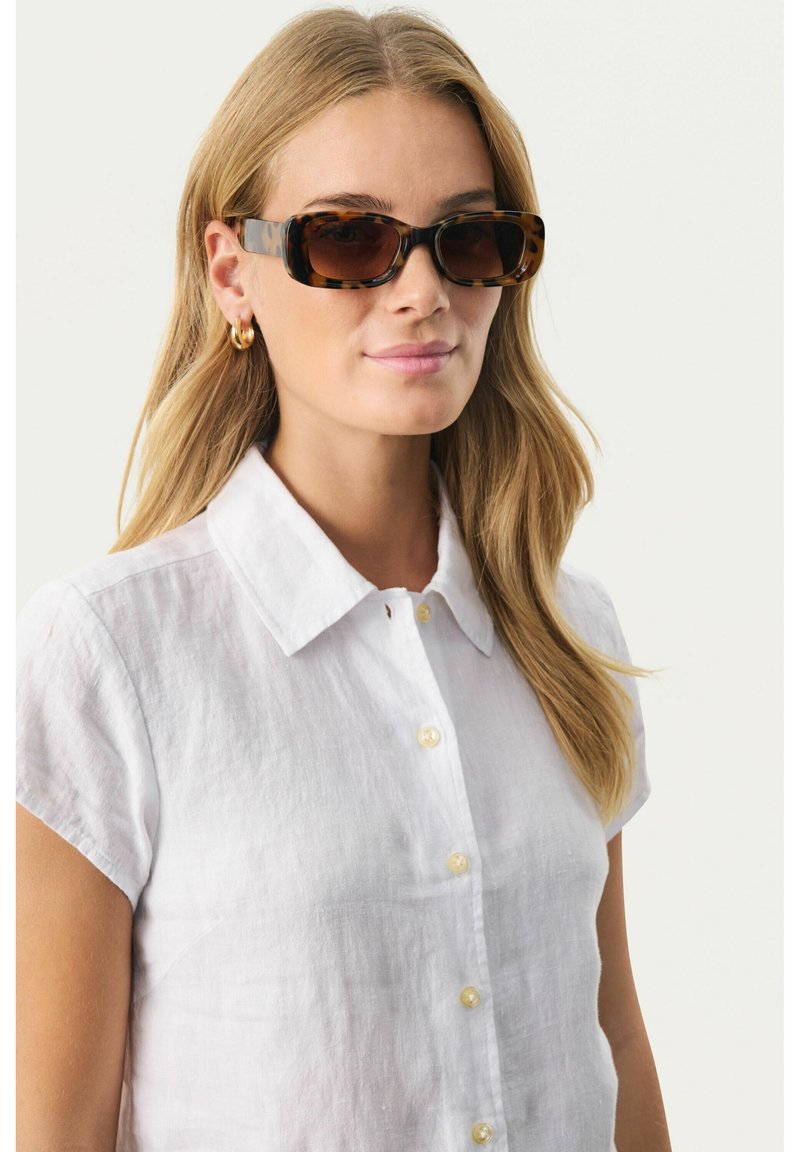 Lunettes de soleil écaille de tortue avec une forme rectangulaire, une teinte neutre et un motif subtil. Le modèle porte une chemise en lin blanche à col avec des boutons.