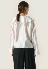 Blusa de satén blanco con un amplio cuello rizado y mangas largas. La tela tiene una textura suave y un brillo sutil. Combinada con pantalones rayados negros.