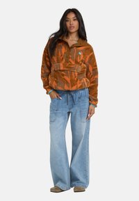 Bruine en oranje fleece pullover met een kraag, halve knoopsluiting en een voorzak; gecombineerd met losse lichtblauwe jeans.