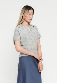 Donna con lunghi capelli ombre, che indossa un maglione tricot grigio a maniche corte e una gonna di raso blu, in piedi con le mani unite davanti.