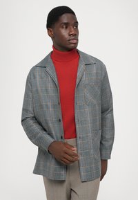 Hall toogri riidest jakk oranzide ja siniste aktsentidega, millel on rinna tasku ja nupuga sulgemine, koos punase kõrge kaelusega sviitriga.