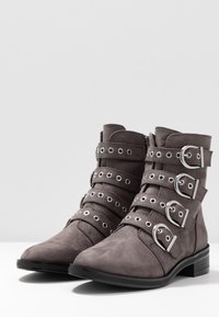 Botas de tobillo de ante sintético gris con múltiples correas, herrajes de tono plateado, detalles en forma de anillo y un tacón ancho. Textura suave y punta redondeada.