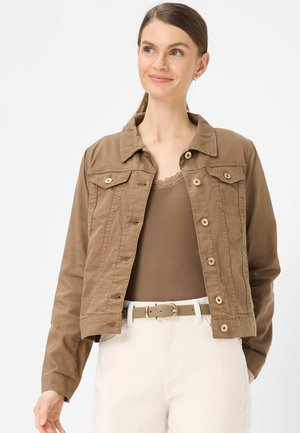 Femme portant une veste marron boutonnée par-dessus un haut marron avec bordure en dentelle, un pantalon blanc et une ceinture beige, debout devant un fond neutre.