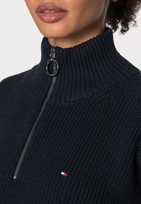 Pull à col montant bleu marine avec texture côtelée, doté d'une fermeture éclair sur le devant et d'un petit logo rouge, blanc et bleu sur la poitrine.