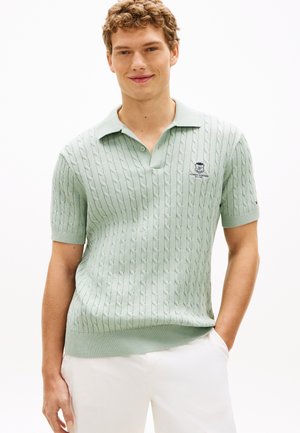 Jeune homme aux cheveux bouclés portant un polo à manches courtes en maille torsadée vert clair et un pantalon blanc, les mains détendues le long du corps.