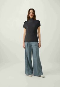 Must have lühikeste varrukatega must topp, millel on kõrge kaelus, kombineeritud helesiniste laia säärtega teksadega. Mudel seisab ühtlase tausta ees.